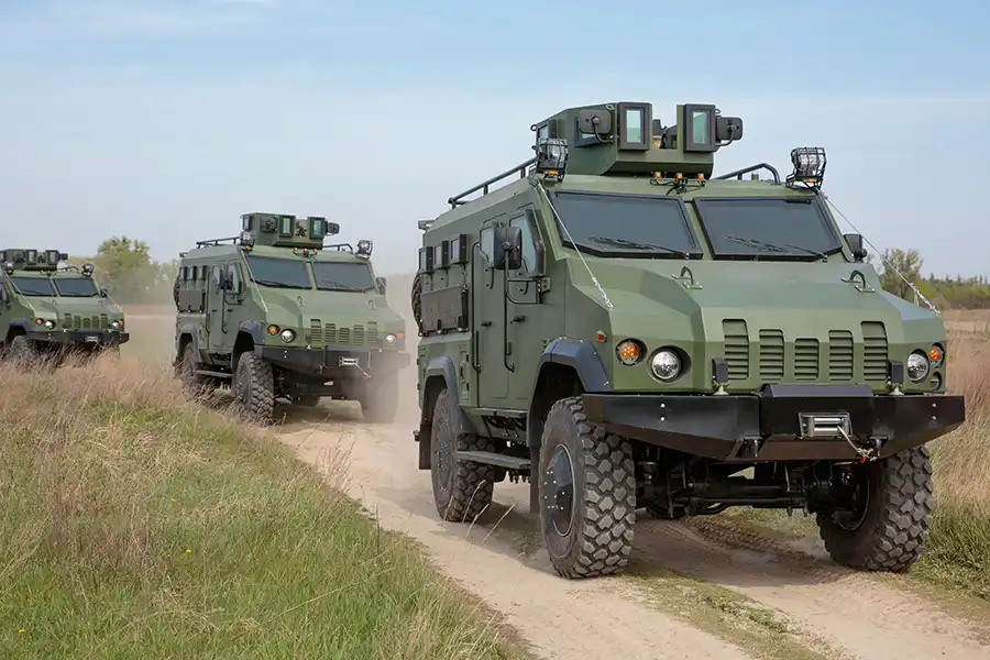 Row of green military armored vehicles with heavy-duty tires driving along a dirt field road under blue sky at AUSA 2025